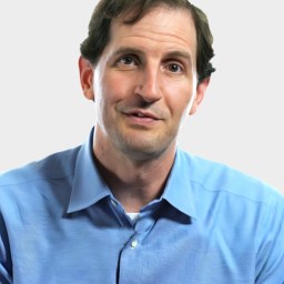 A man with short brown hair wearing a light blue button-up shirt poses against a plain light background.