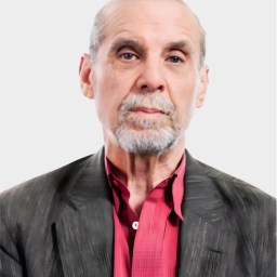 An older man with gray hair and beard wearing a dark jacket over a red shirt, looking straight at the camera against a plain light background.