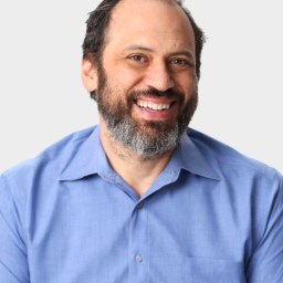 A man with dark hair and a beard, wearing a light blue collared shirt, smiles at the camera against a plain light background.