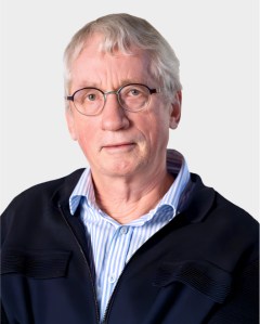 Older man with short gray hair and glasses, wearing a striped shirt and dark jacket, looking at the camera against a plain light background.