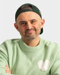 A man with a beard wearing a green sweater with a white heart and a backwards green cap, smiling with arms crossed against a plain background.
