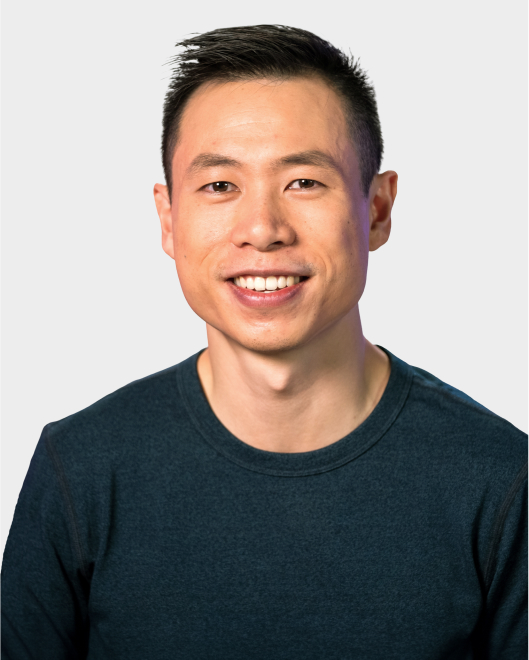 A man with short black hair, wearing a dark blue crew neck shirt, smiling at the camera against a plain light background.
