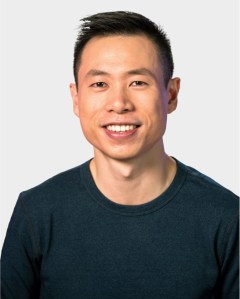 A man with short black hair, wearing a dark blue crew neck shirt, smiling at the camera against a plain light background.
