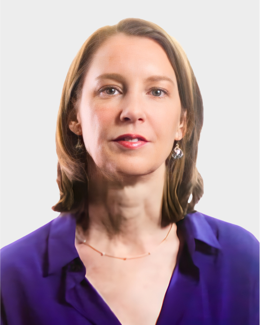 A woman with shoulder-length brown hair, wearing a purple blouse, drop earrings, and a delicate necklace, poses against a plain light background.