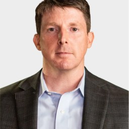 A man with short brown hair wearing a gray suit jacket and light blue shirt, facing forward with a neutral expression against a plain background.