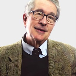 An older man with gray hair and glasses is wearing a brown herringbone jacket, a collared shirt, and a dark sweater, posing against a plain light background.
