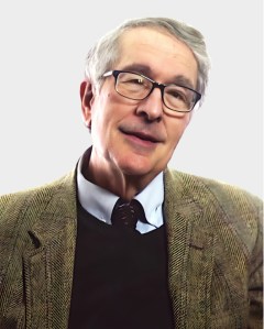 An older man with gray hair and glasses is wearing a brown herringbone jacket, a collared shirt, and a dark sweater, posing against a plain light background.