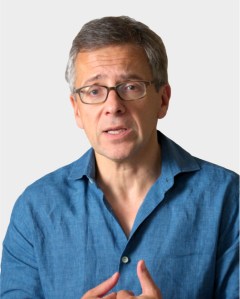 A middle-aged man with glasses and gray hair, wearing a blue shirt, speaks while gesturing with his hands against a plain background.