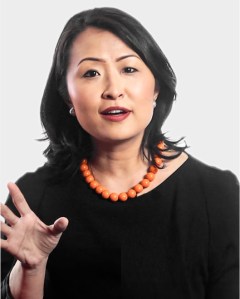 A woman with medium-length black hair wears a black top and an orange beaded necklace, gesturing with her left hand while speaking against a plain light background.