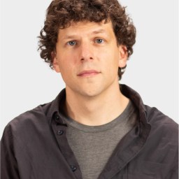 A man with curly brown hair wearing a dark button-up shirt over a gray t-shirt, looking directly at the camera against a plain light background.