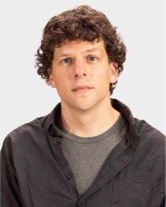 A man with curly brown hair wearing a dark button-up shirt over a gray t-shirt, looking directly at the camera against a plain light background.