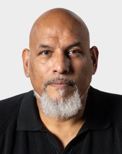 A middle-aged man with a bald head, gray goatee, and mustache, wearing a black collared shirt, facing forward against a plain background.