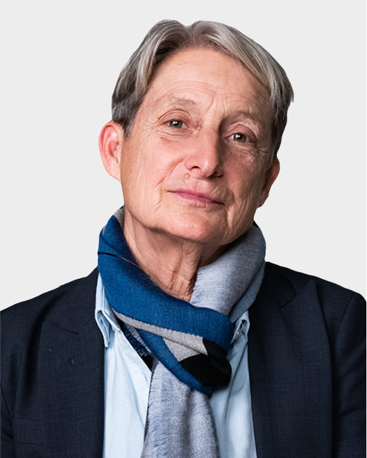 An older person with short gray hair, wearing a dark blazer, light blue shirt, and a blue and gray scarf, poses against a plain light background.