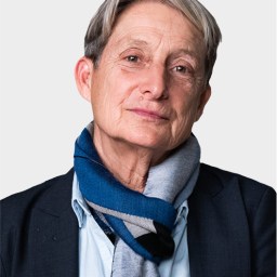 An older person with short gray hair, wearing a dark blazer, light blue shirt, and a blue and gray scarf, poses against a plain light background.