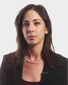 A woman with long brown hair, wearing a dark blazer, maroon top, and a beaded necklace, poses against a plain white background.