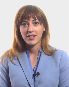 A woman with light brown hair wearing a light blue blazer and a blue shirt, speaking and facing the camera against a plain white background.