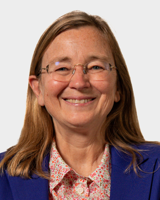 A woman with long light brown hair, glasses, and a blue blazer over a floral shirt smiles at the camera against a plain background.