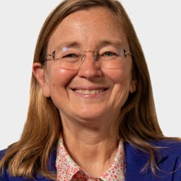 A woman with long light brown hair, glasses, and a blue blazer over a floral shirt smiles at the camera against a plain background.