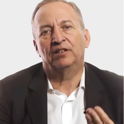 A middle-aged man in a dark suit and white shirt speaks, gesturing with his hand, against a plain light background.
