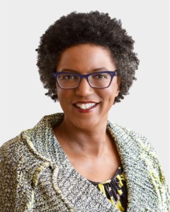 A woman with short curly hair and glasses smiles at the camera. She is wearing a patterned jacket over a patterned top, in front of a plain light background.