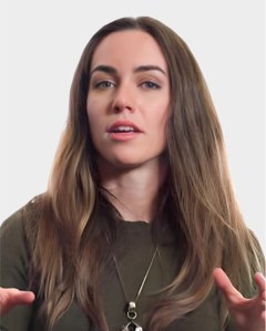 A woman with long brown hair and a neutral expression gestures with her hands. She is wearing a dark top and a pendant necklace against a plain background.