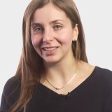 A woman with long brown hair, wearing a black top and a necklace, looks at the camera and smiles against a plain light background.