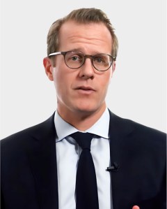A man wearing glasses, a dark suit, white shirt, and dark tie stands against a plain light background, looking directly at the camera.