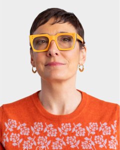 Person with short dark hair wearing large yellow glasses, gold hoop earrings, and an orange sweater with a white floral pattern, facing forward against a plain light background.