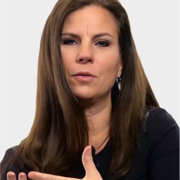 Woman with long brown hair wearing a black top, gesturing with her hand while speaking, against a plain light background.