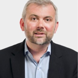 A man with short gray hair and a beard, wearing a dark blazer over a light blue shirt, poses against a plain light background.
