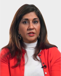 Woman with long brown hair wearing a red blazer and white top, seated against a plain light background, looking directly at the camera.