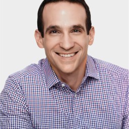 A man with dark hair, wearing a purple and white checkered shirt, smiles at the camera against a plain white background.