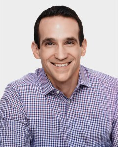 A man with dark hair, wearing a purple and white checkered shirt, smiles at the camera against a plain white background.