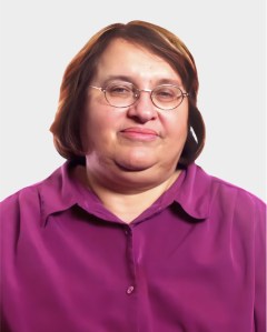 A middle-aged woman with short brown hair, glasses, and a purple button-up shirt is looking at the camera against a plain light background.