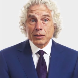 A man with curly gray hair wearing a blue suit, white shirt, and dark tie poses against a plain light background.