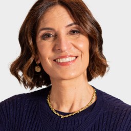A woman with shoulder-length brown hair, wearing a navy sweater, gold necklace, and earrings, smiles at the camera against a plain light background.