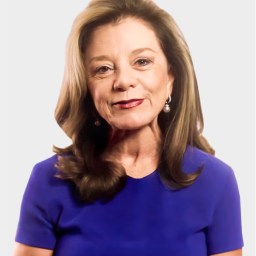 A woman with shoulder-length brown hair, wearing a blue top and pearl earrings, stands against a plain light background.