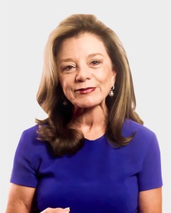 A woman with shoulder-length brown hair, wearing a blue top and pearl earrings, stands against a plain light background.