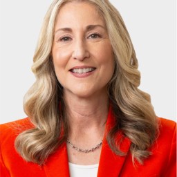 A woman with long, wavy blonde hair wearing an orange blazer, white top, and silver necklace, smiling at the camera against a plain light background.