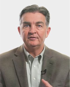 Middle-aged man wearing a gray blazer and light shirt, looking at the camera with a neutral expression, with a microphone clipped to his jacket.