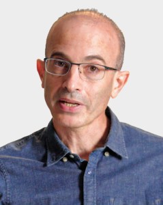 A middle-aged person with glasses and a receding hairline, wearing a blue button-up shirt, is speaking against a plain light background.