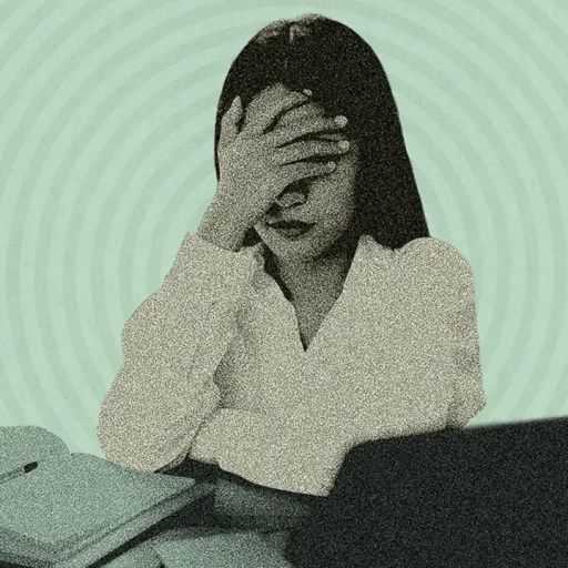 A person sitting at a desk with a laptop and papers, covering their face with one hand, suggesting stress or frustration.