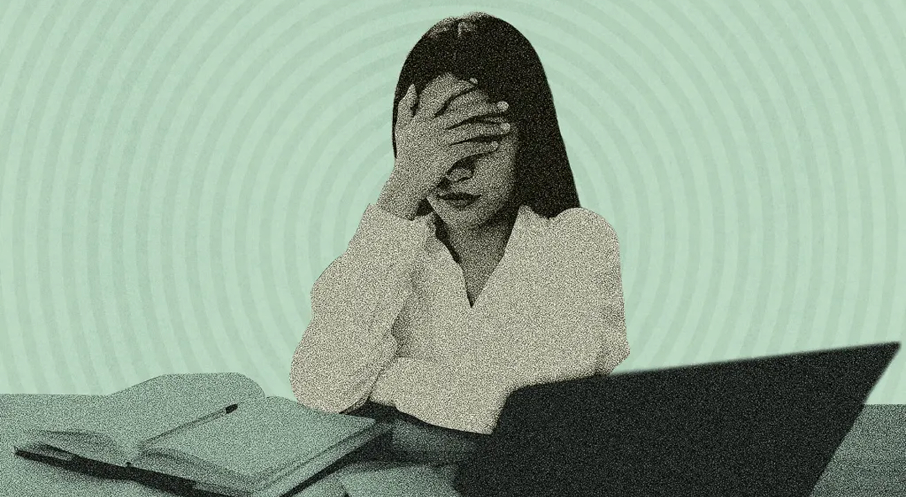 A person sitting at a desk with a laptop and papers, covering their face with one hand, suggesting stress or frustration.