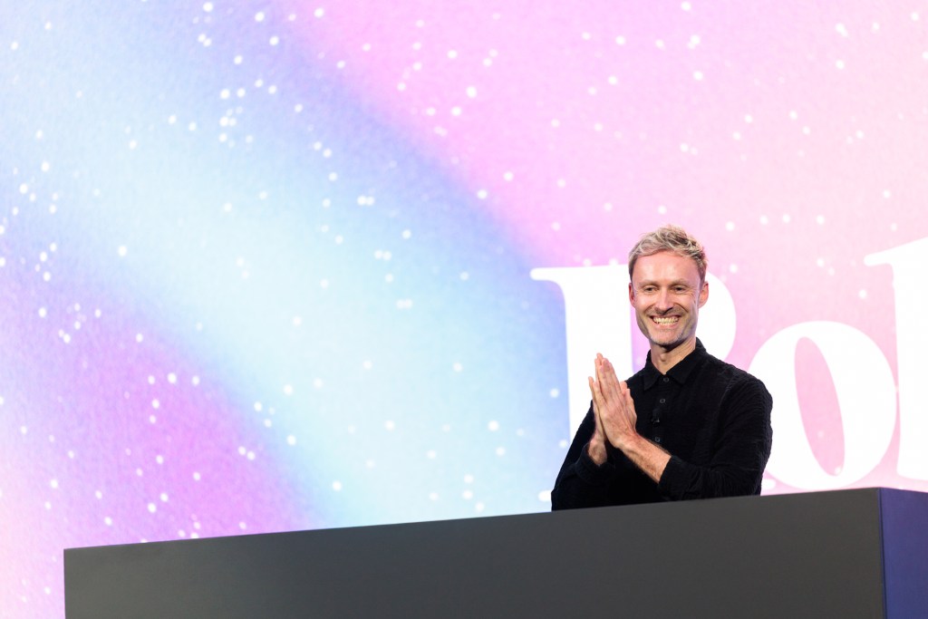 A man stands behind a podium, smiling with his hands clasped, against a colorful background with white specks and large text partially visible.