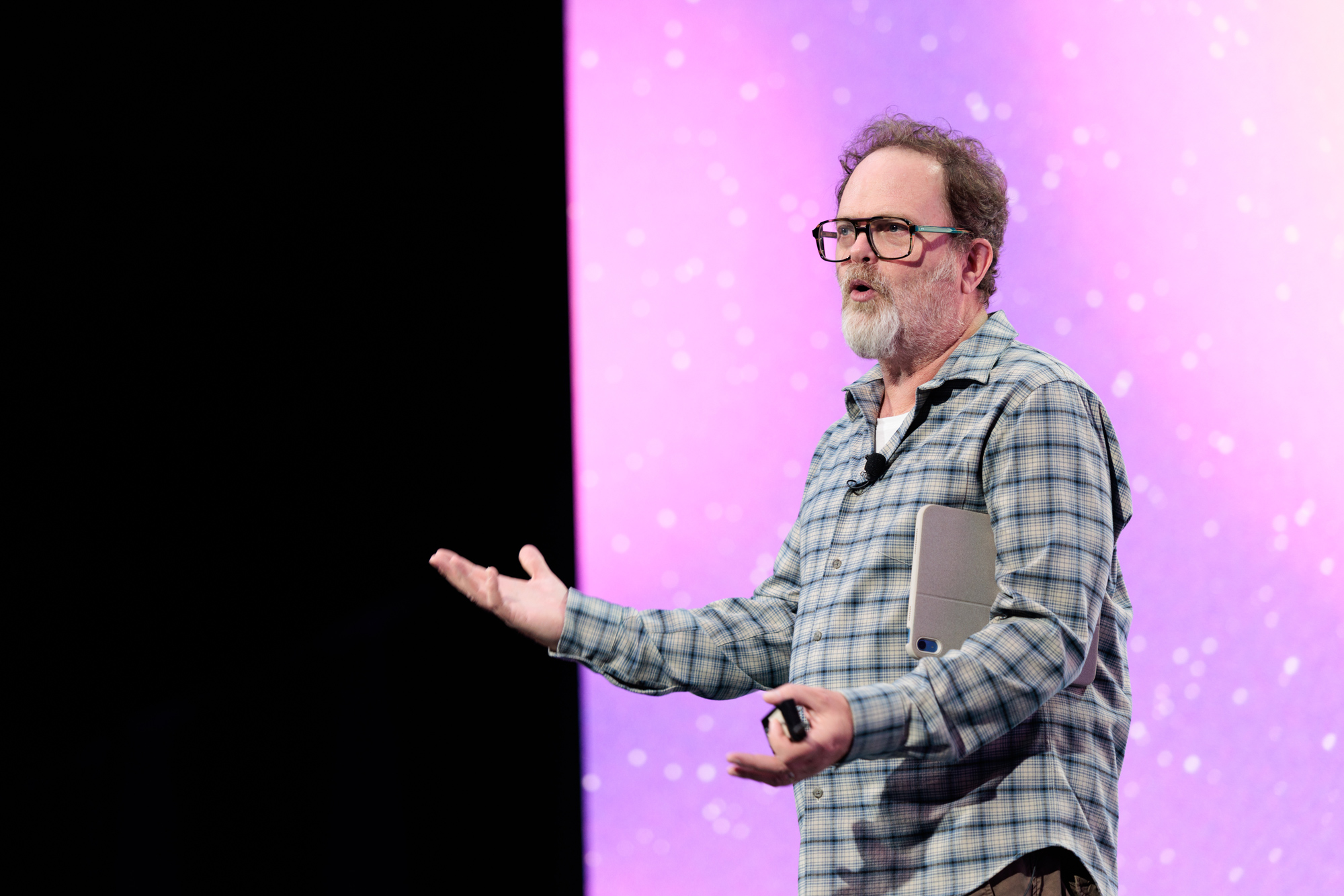 A man in a plaid shirt speaks on stage with a tablet under his arm, gesturing with his hands, in front of a purple backdrop.