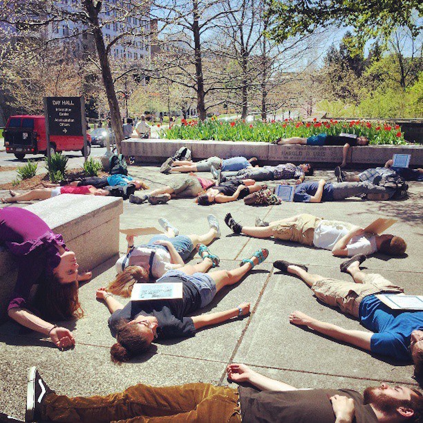 A group of people are lying on the ground in an outdoor plaza on a sunny day, some holding signs about alien life, in what appears to be a public demonstration or protest.
