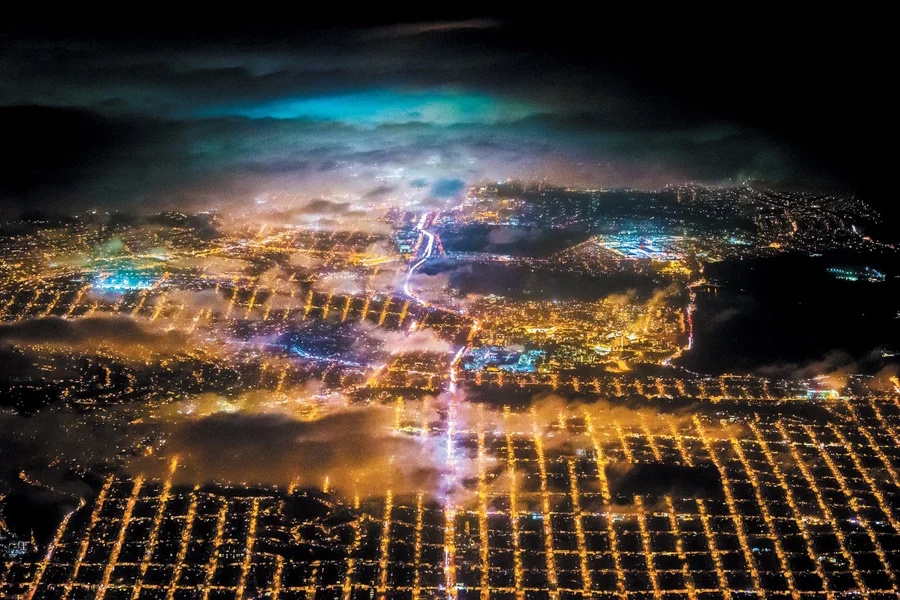 Aerial night view of a city grid with illuminated streets and buildings, partially covered by clouds and mist, under a dark sky—showcasing how solar power at night enhances urban lighting.