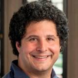 A man with curly dark hair and a blue collared shirt smiles at the camera; blurred background with windows and wood paneling.