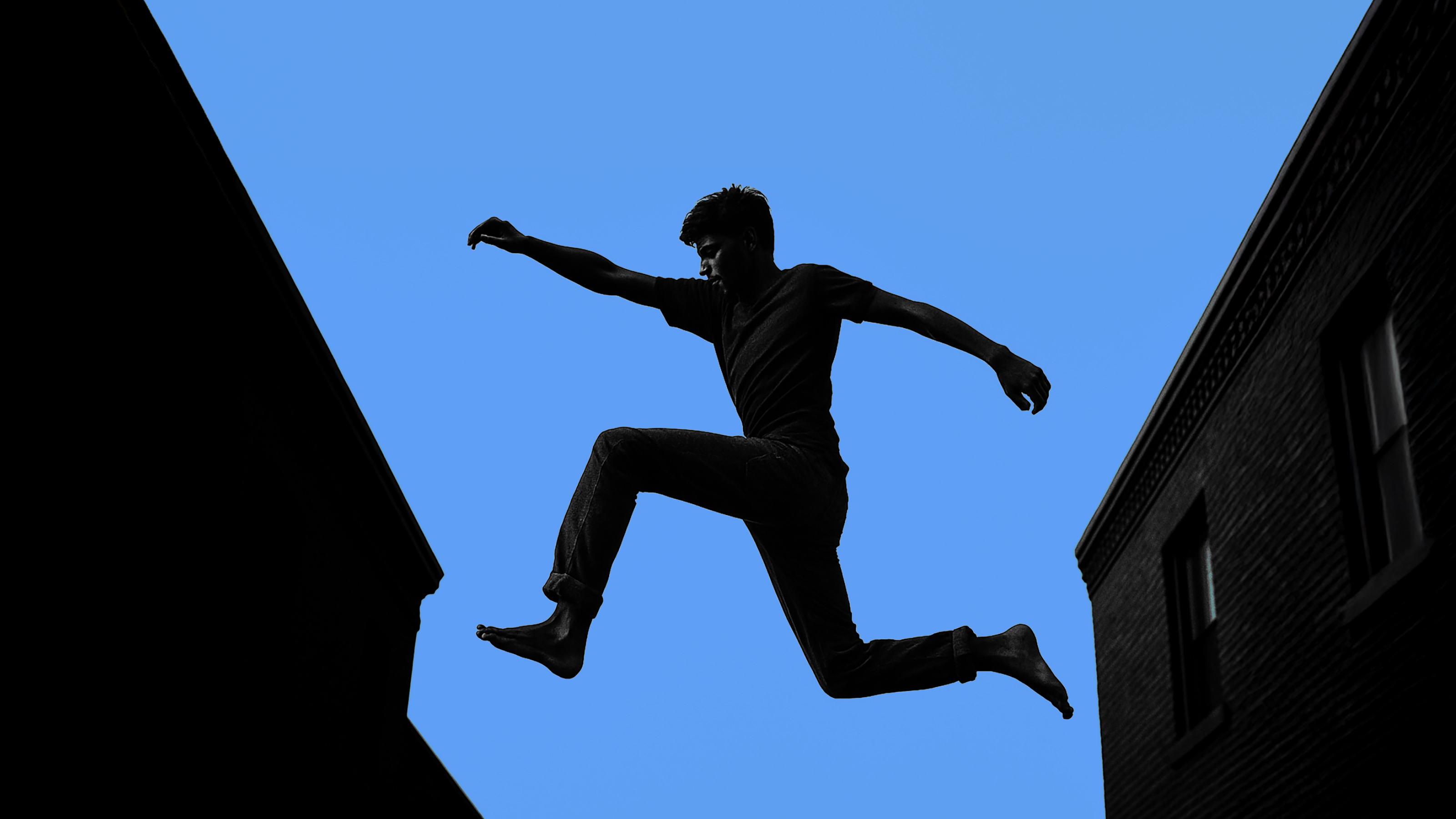 A person is leaping between two buildings against a clear blue sky.