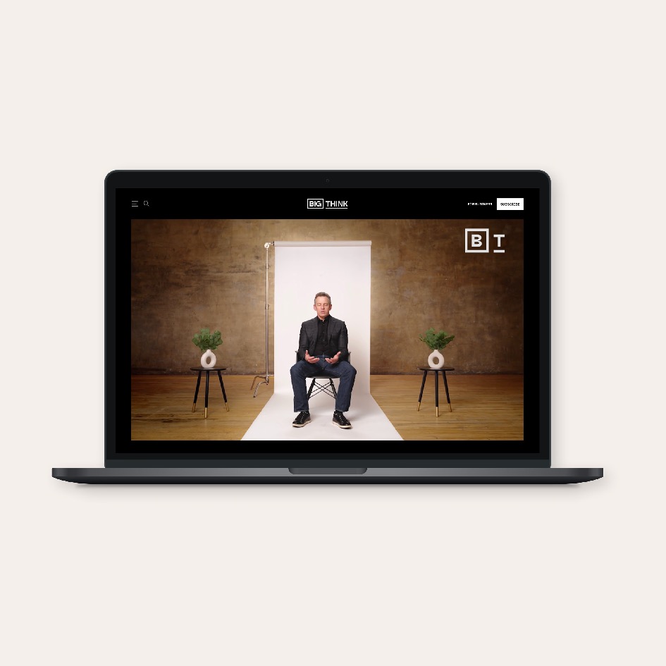 A laptop displaying a video of a man sitting on a stool in a studio with wooden flooring, plants on stands, and a white backdrop behind him.